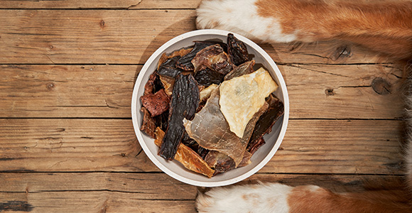 a bowl of various dog treats on a wooden surface with the paws of a dog visible nearby perfect for six healthy snacks for pets